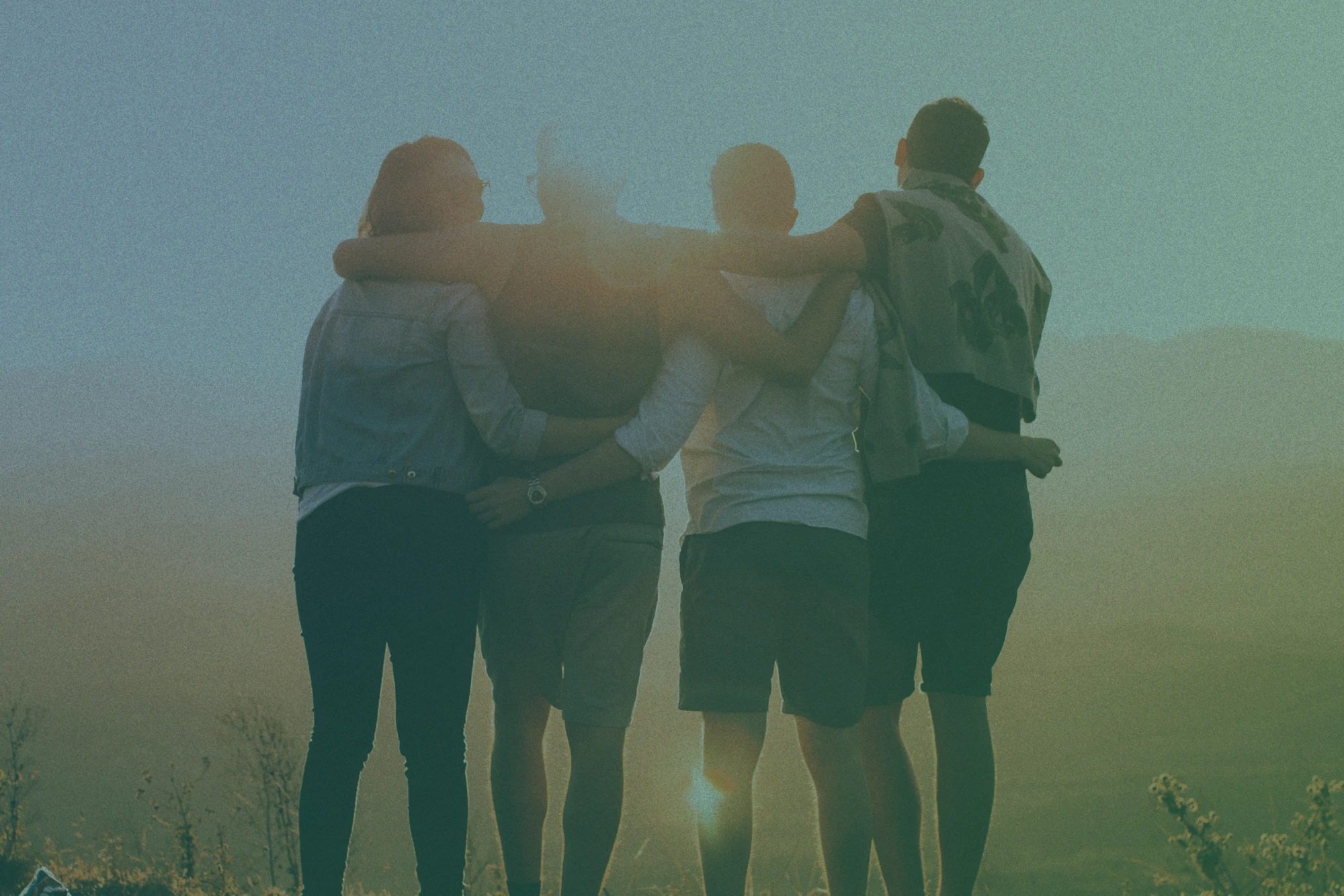 A group of friends in nature, viewed from behind, with arms around each others' shoulders